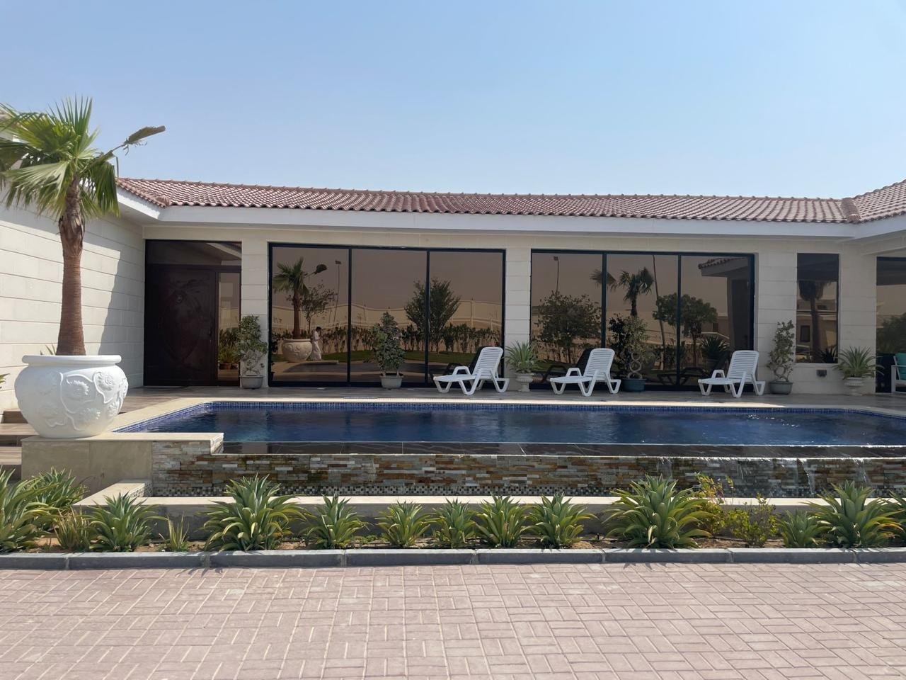 The estate’s pool pavilion — a single-storey colonnade in tinted glass facing a long pool, shaded terraces and mature palms against open desert sky.